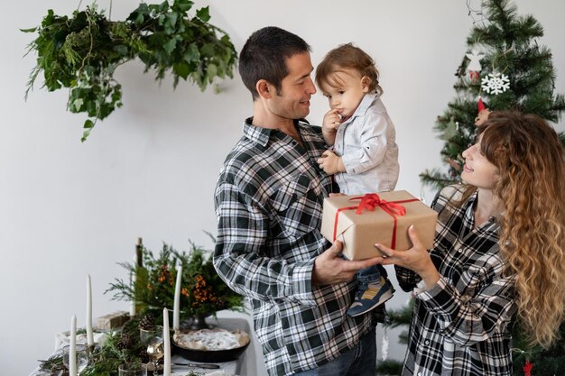 Navidad en casa una madre de familia le da un regalo a un hijo pequeño sentado en los brazos de su padre en la sala de estar decorada con un árbol de Navidad y una mesa con velas Sorpresa de Año Nuevo para el niño