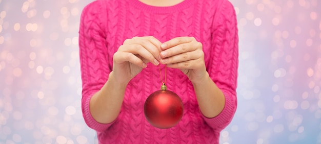 Navidad, decoración, vacaciones y concepto de la gente: cerca de una mujer con suéter rosa sosteniendo una bola de Navidad sobre cuarzo rosa y fondo de luces de serenidad