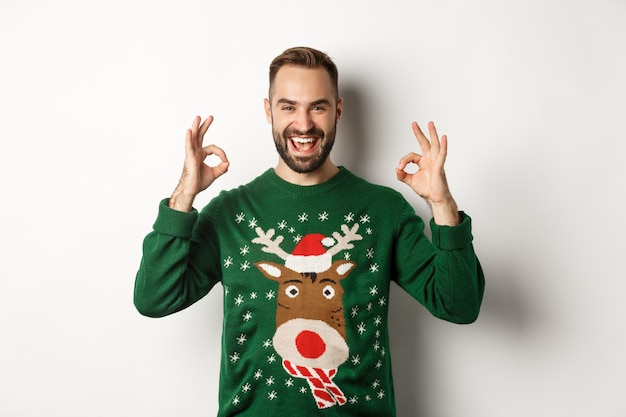 Navidad, fiestas y celebraciones. Chico guapo satisfecho en suéter, mostrando signos de bien y sonriendo, alaba algo grande, de pie sobre fondo blanco.