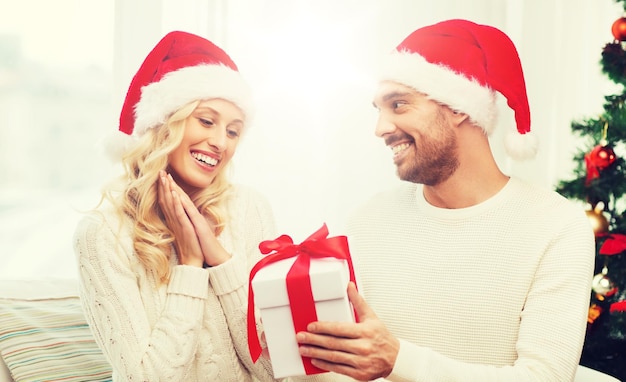 Navidad, fiestas y concepto de personas - pareja feliz con sombreros de Santa con caja de regalos sentada en el sofá en casa