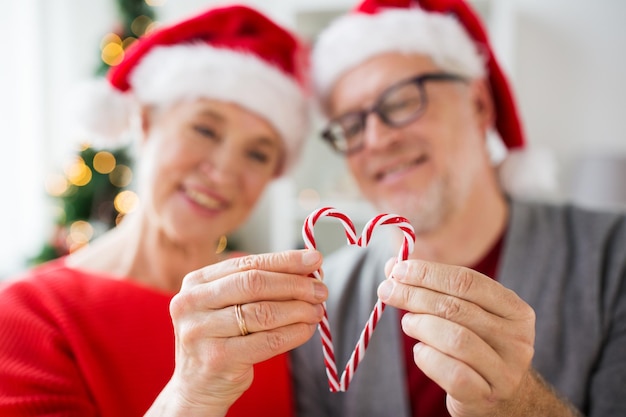 Navidad, fiestas y concepto de personas - primer plano de una feliz pareja de ancianos sonrientes con sombreros de Papá Noel con corazones de bastones de caramelo en casa