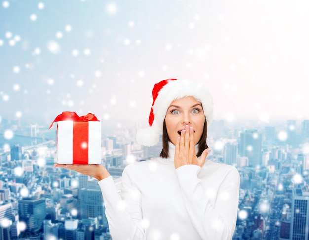navidad, invierno, felicidad, vacaciones y concepto de la gente - mujer sonriente con sombrero de ayudante de santa con caja de regalo sobre fondo de ciudad nevada