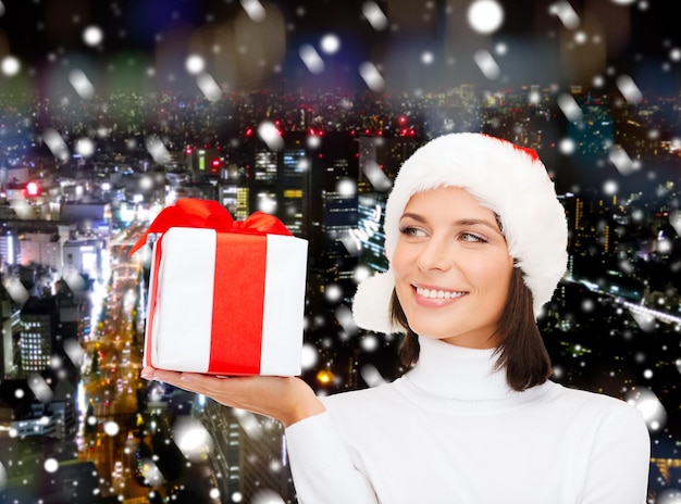 navidad, invierno, felicidad, vacaciones y concepto de la gente - mujer sonriente con sombrero de ayudante de santa con caja de regalo sobre el fondo de la ciudad de noche nevada