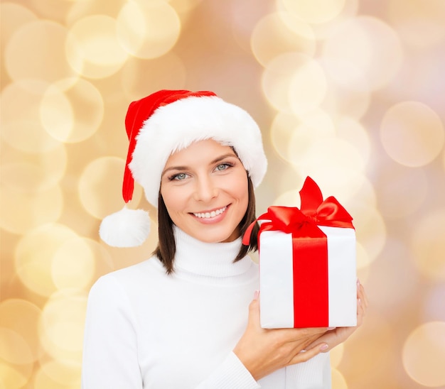 navidad, invierno, felicidad, vacaciones y concepto de la gente - mujer sonriente con sombrero de ayudante de santa con caja de regalo sobre fondo de luces beige