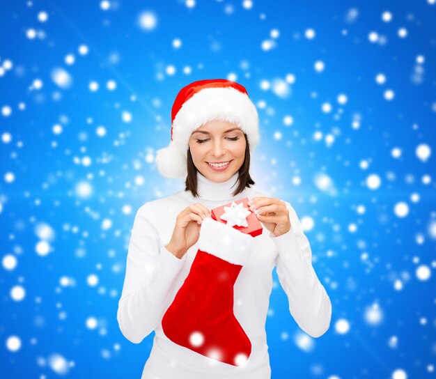 navidad, invierno, felicidad, vacaciones y concepto de la gente - mujer sonriente con sombrero de ayudante de santa con una pequeña caja de regalo y calcetín sobre fondo azul nevado