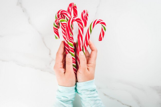 Navidad, invierno Fiestas, dulces, golosinas. Manos de niña sosteniendo el bastón de caramelo tradicional en forma de un ramo. Mesa de mármol blanco, vista superior, copyspace