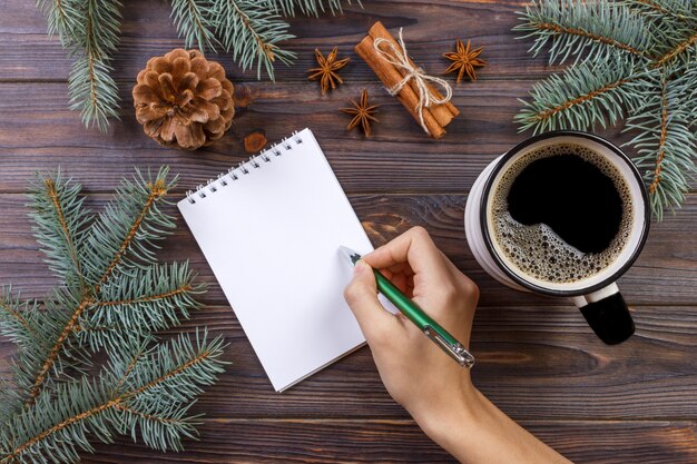 Navidad de manos femeninas escribiendo en cuaderno abierto sobre mesa de madera rústica cubierta con decoración navideña, vista superior, endecha plana