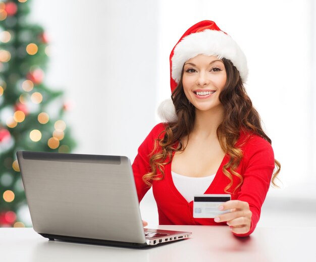 Navidad, Navidad, concepto de compras online - mujer con sombrero de ayudante de santa con ordenador portátil y tarjeta de crédito