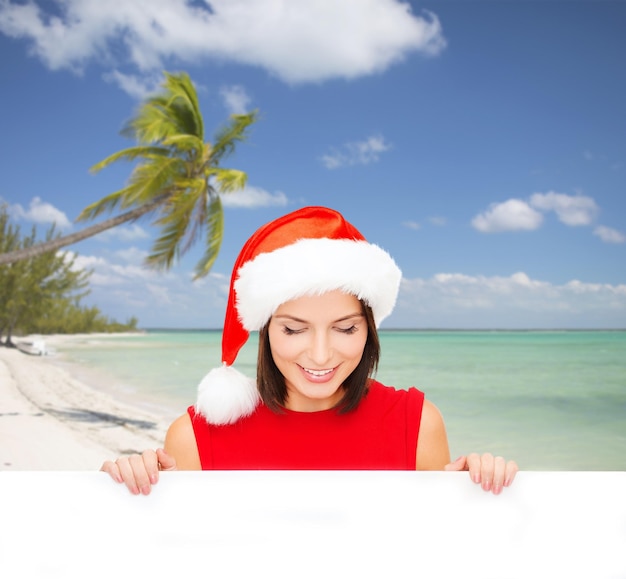 navidad, navidad, gente, publicidad y concepto de venta - mujer feliz con sombrero de ayudante de santa con pizarra en blanco sobre fondo de playa tropical