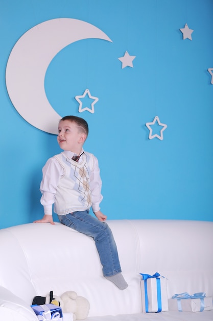 Navidad. niño en un sofá blanco con caja de regalos de Navidad. pared azul con una luna blanca en una pared.