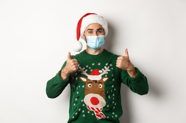 Navidad durante la pandemia, concepto covid-19. Hombre satisfecho con gorro de Papá Noel y máscara médica mostrando el pulgar hacia arriba en señal de aprobación, alabando algo, de pie sobre fondo blanco.