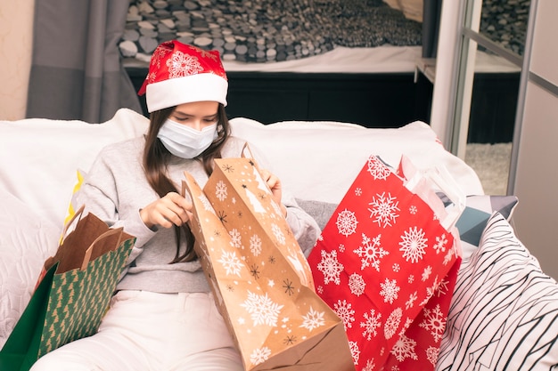 Navidad durante la pandemia de coronavirus covid 19, mujer joven con máscara médica en casa mira los regalos de Año Nuevo en bolsas