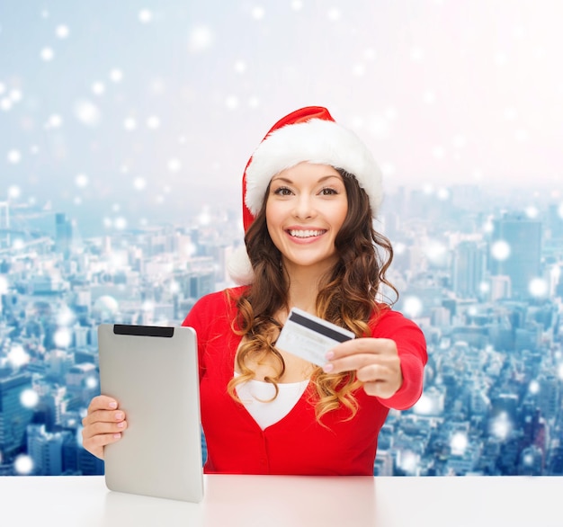 Navidad, tecnología, compras y concepto de personas: mujer sonriente con sombrero de ayudante de santa con computadora de tablet pc y tarjeta de crédito sobre fondo de ciudad nevada