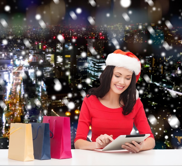 Navidad, tecnología y concepto de personas: mujer sonriente con sombrero de ayudante de santa con bolsas de compras y computadora de tablet pc sobre el fondo de la ciudad de noche nevada