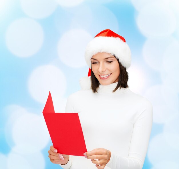 navidad, vacaciones, celebración, saludo y concepto de la gente - mujer sonriente con sombrero de ayudante de santa con tarjeta de saludo sobre fondo de luces azules