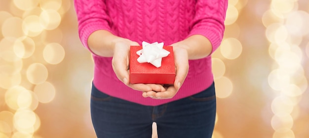 Navidad, vacaciones y concepto de la gente: cerca de una mujer con suéter rosa sosteniendo una caja de regalo sobre fondo de luces beige