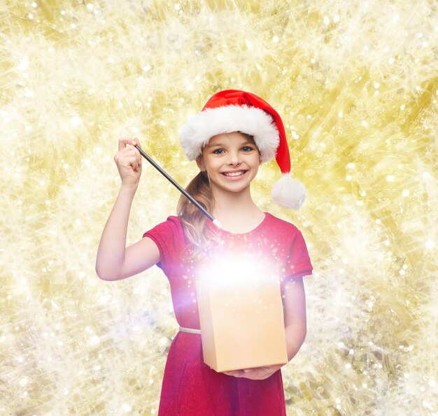 navidad, vacaciones, felicidad y concepto de la gente - chica sonriente con sombrero de ayudante de santa con caja de regalo y varita mágica sobre fondo de luces amarillas