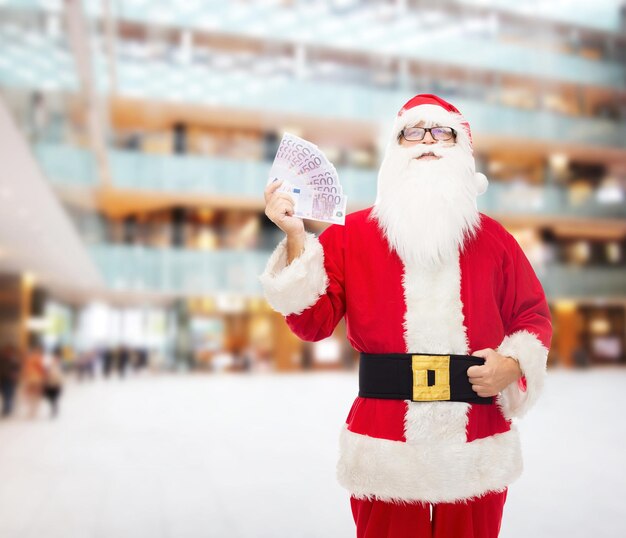 navidad, vacaciones, ganar, moneda y concepto de personas - hombre disfrazado de santa claus con dinero en euros sobre el fondo del centro comercial