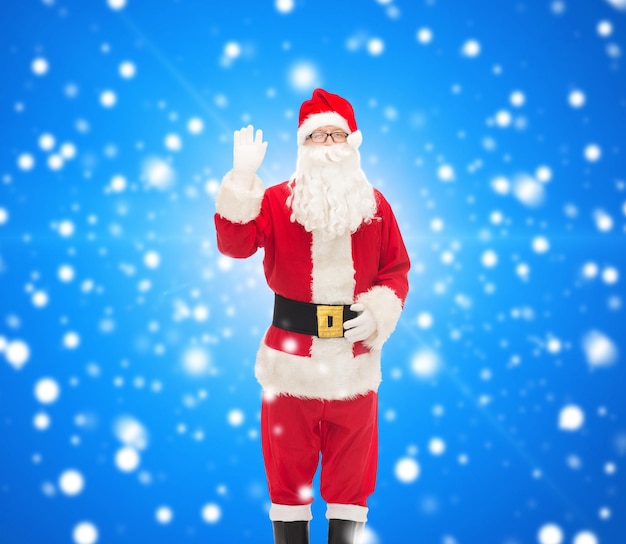 navidad, vacaciones, gesto y concepto de la gente - hombre disfrazado de santa claus agitando la mano sobre fondo azul nevado