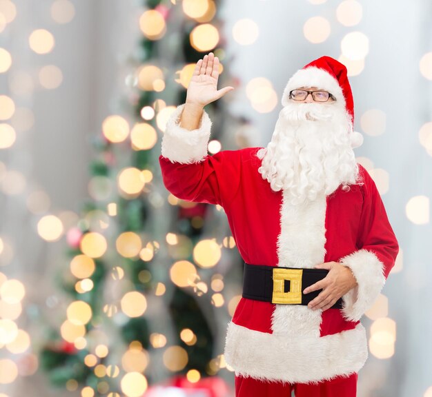 navidad, vacaciones, gesto y concepto de la gente - hombre disfrazado de santa claus agitando la mano sobre el fondo de las luces del árbol