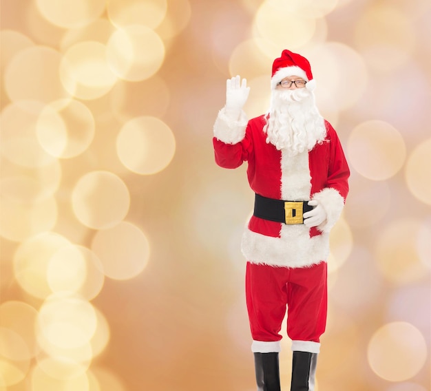 navidad, vacaciones, gesto y concepto de la gente - hombre disfrazado de santa claus agitando la mano sobre fondo de luces beige