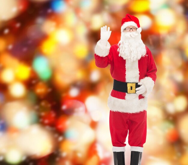 navidad, vacaciones, gesto y concepto de la gente - hombre disfrazado de santa claus agitando la mano sobre fondo de luces rojas