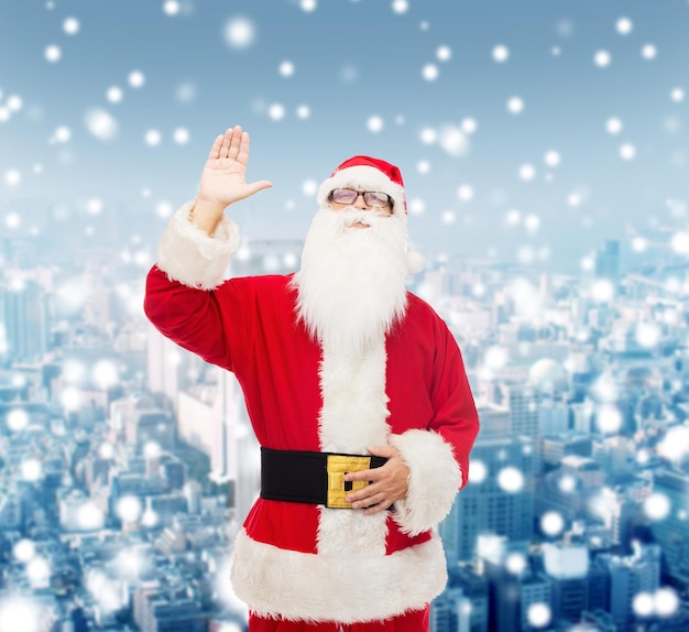 navidad, vacaciones, gesto y concepto de la gente - hombre disfrazado de santa claus agitando la mano sobre el fondo nevado de la ciudad