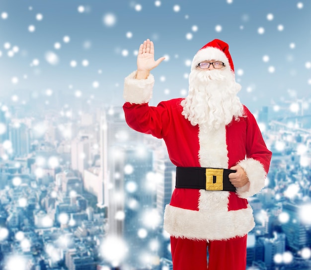 navidad, vacaciones, gesto y concepto de la gente - hombre disfrazado de santa claus agitando la mano sobre el fondo nevado de la ciudad