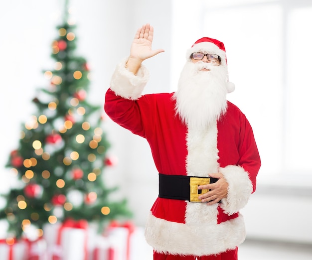 navidad, vacaciones, gesto y concepto de la gente - hombre disfrazado de santa claus agitando la mano sobre la sala de estar con fondo de árbol