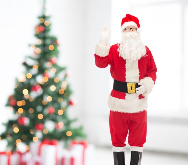 navidad, vacaciones, gesto y concepto de la gente - hombre disfrazado de santa claus agitando la mano sobre la sala de estar y el fondo del árbol