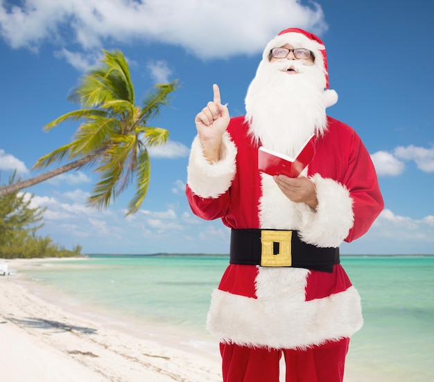 navidad, vacaciones, gesto y concepto de la gente - hombre disfrazado de santa claus con bloc de notas apuntando con el dedo hacia arriba sobre el fondo de la playa tropical