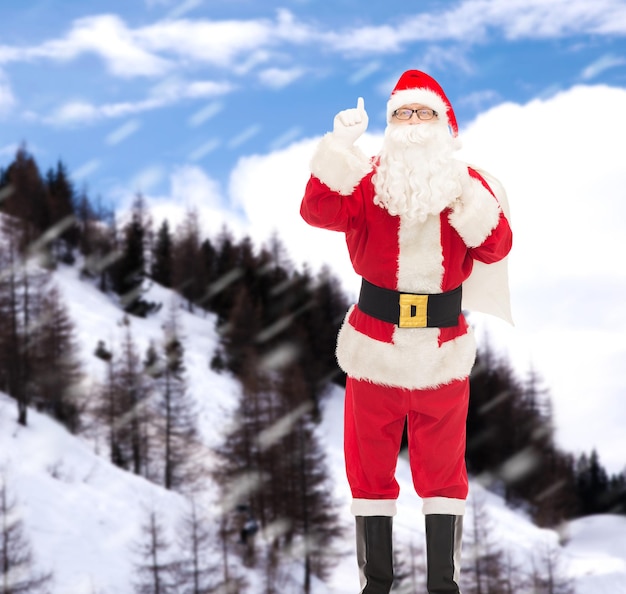 Foto navidad, vacaciones, gesto y concepto de la gente - hombre disfrazado de santa claus con una bolsa apuntando con el dedo hacia arriba sobre el fondo de las montañas nevadas