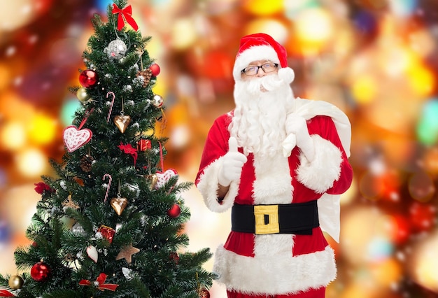 navidad, vacaciones, gesto y concepto de la gente - hombre disfrazado de santa claus con bolsa y árbol de navidad mostrando los pulgares hacia arriba sobre fondo de luces rojas