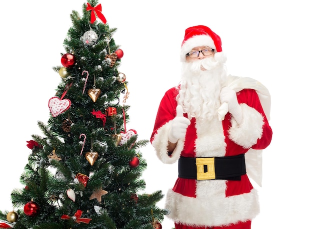 navidad, vacaciones, gesto y concepto de la gente - hombre disfrazado de santa claus con bolsa y árbol de navidad mostrando los pulgares hacia arriba