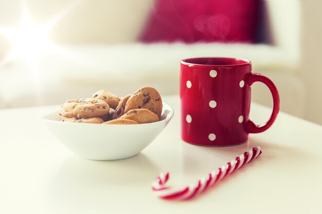 Foto navidad, vacaciones, invierno y concepto de celebración - primer plano de galletas de avena, dulces de caña de azúcar y taza roja en la mesa en casa