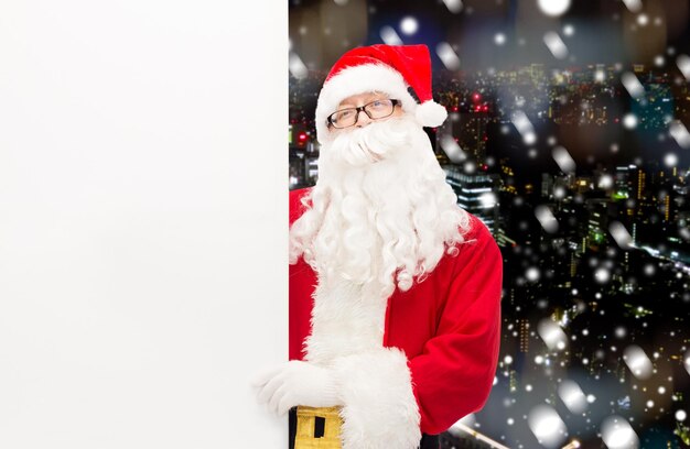 navidad, vacaciones, publicidad y concepto de la gente - hombre disfrazado de santa claus con cartel blanco en blanco sobre el fondo de la ciudad de noche nevada