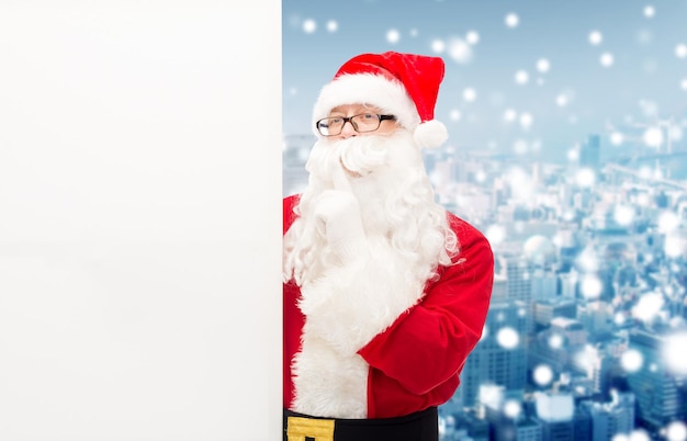 navidad, vacaciones, publicidad y concepto de la gente - hombre disfrazado de santa claus con cartelera blanca en blanco haciendo un gesto de hust sobre el fondo nevado de la ciudad