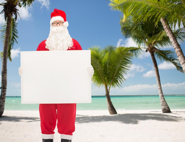 navidad, vacaciones, publicidad y concepto de la gente - hombre disfrazado de santa claus con cartelera blanca en blanco sobre fondo de playa tropical