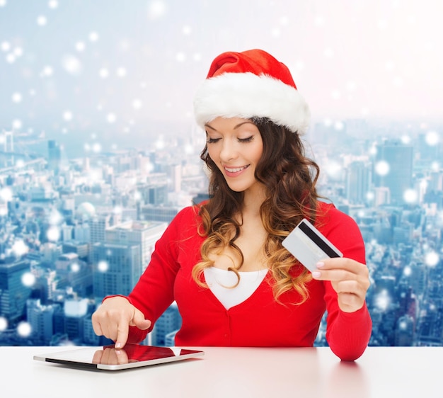 navidad, vacaciones, tecnología y concepto de compras - mujer sonriente con sombrero de ayudante de santa con tarjeta de crédito y computadora de tablet pc sobre fondo de ciudad nevada