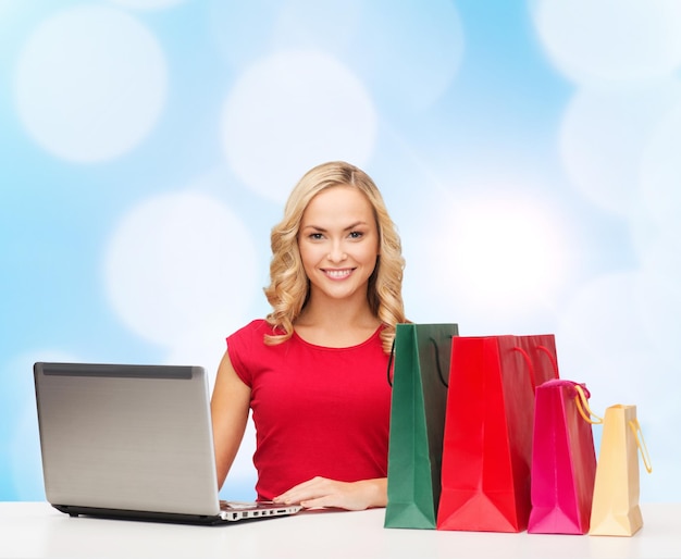 navidad, vacaciones, tecnología, publicidad y concepto de personas - mujer sonriente con pantalones rojo en blanco con bolsas de compras y computadora portátil sobre fondo de luces azules