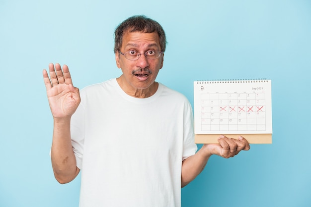 Índio sênior segurando um calendário isolado em um fundo azul surpreso e chocado.