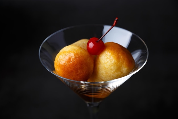 Neapolitanischer Rum Baba oder Baba au Rum in einem Martini-Glas mit einer Cocktailkirsche auf schwarzem Hintergrund Kleine Hefekuchen, die in Rumsirup getränkt sind Traditionelles italienisches Gebäck Closeup selektiver Fokus