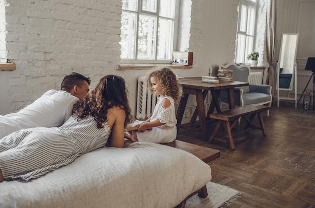 Foto neben dem bett, auf dem papa und mama liegen, sitzt ein kleines lockiges mädchen. familienwochenende