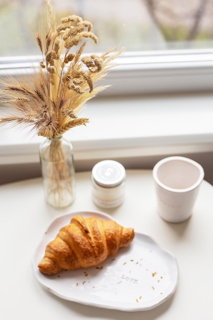 Neben dem Fenster auf dem Tisch stehen ein weißer Teller mit einem Croissant und ein Tonglas mit einer handgefertigten Inschrift