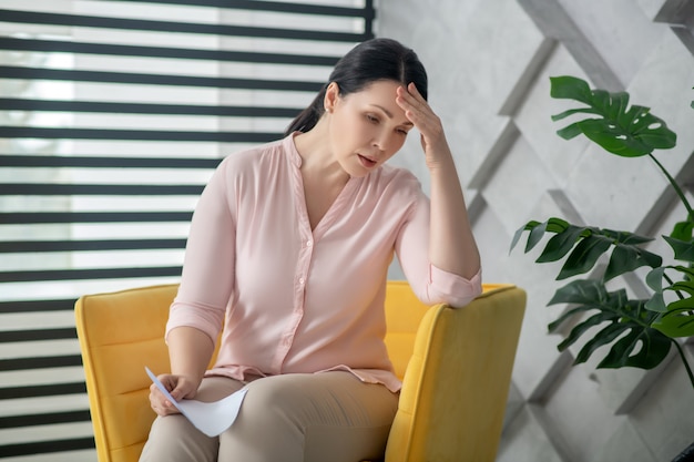 Necesita consulta médica. Triste mujer miserable con una blusa rosa y pantalones, sentada en una silla cerca de la ventana, mirando hacia abajo con la cabeza gacha.