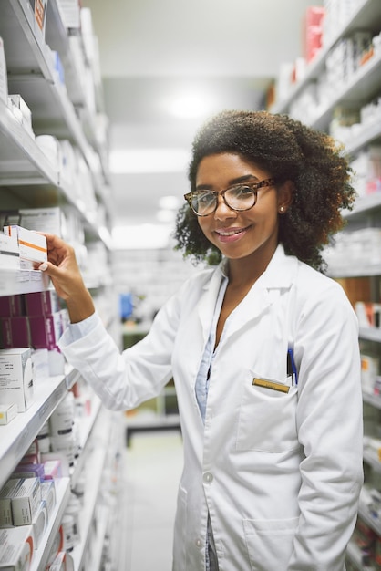 Necesitas algo, lo tenemos Retrato de una joven farmacéutica alegre que pone medicamentos en los estantes mientras mira la cámara en una farmacia