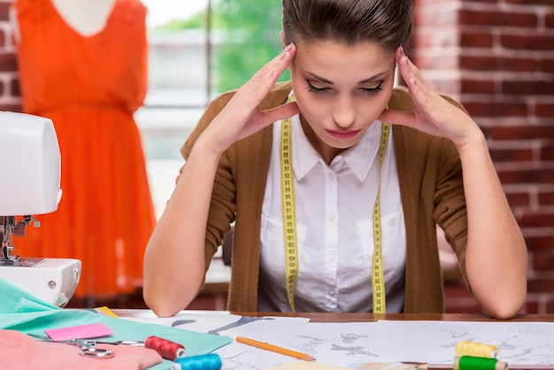 Necesito algunas ideas frescas. Mujer joven cansada sosteniendo la cabeza entre las manos y mirando bocetos de moda mientras está sentada en su lugar de trabajo