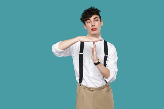 Necesito más tiempo. hombre mostrando gesto de tiempo de espera. Retrato de joven empresario rizado hipster guapo en camisa blanca casual clásica y tirantes de pie. Foto de estudio de interior aislado sobre fondo azul.