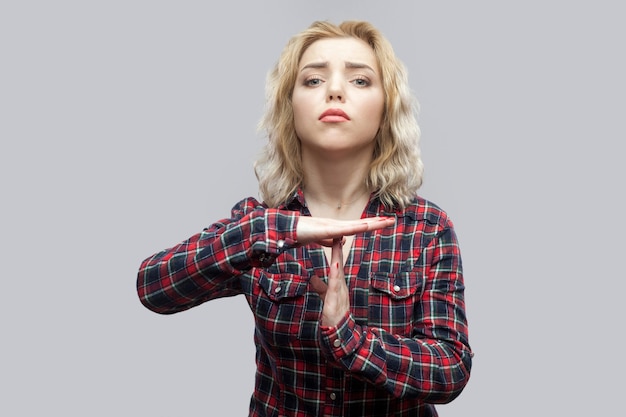 Necesito más tiempo. Retrato de nervos hermosa joven rubia en camisa a cuadros roja casual de pie con signo de tiempo de espera y mirando a cámara. Foto de estudio de interior, aislado sobre fondo gris