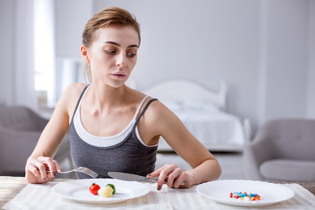 Necesito la medicina. Triste mujer deprimida sentada en la mesa mientras mira las pastillas del plato